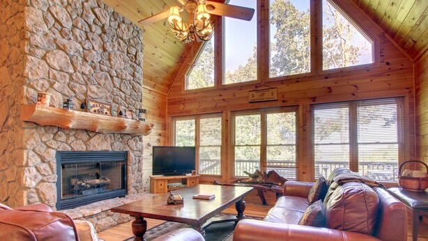 A cozy cabin interior with a stone fireplace, leather seating, and large windows. - Crockett Mountain Lodge Nevaeh Cabin Rentals (Blue Ridge)