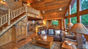 Interior espacioso de una cabaña con una escalera de madera, una sala de estar acogedora con sofá y mesa de centro, y ventanas amplias que ofrecen vistas al exterior.