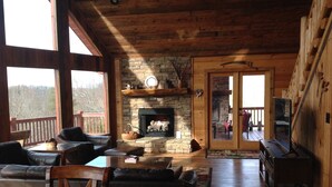 Interior acogedor de una cabaña con chimenea de piedra, muebles de madera y un balcón con vistas a un bosque.