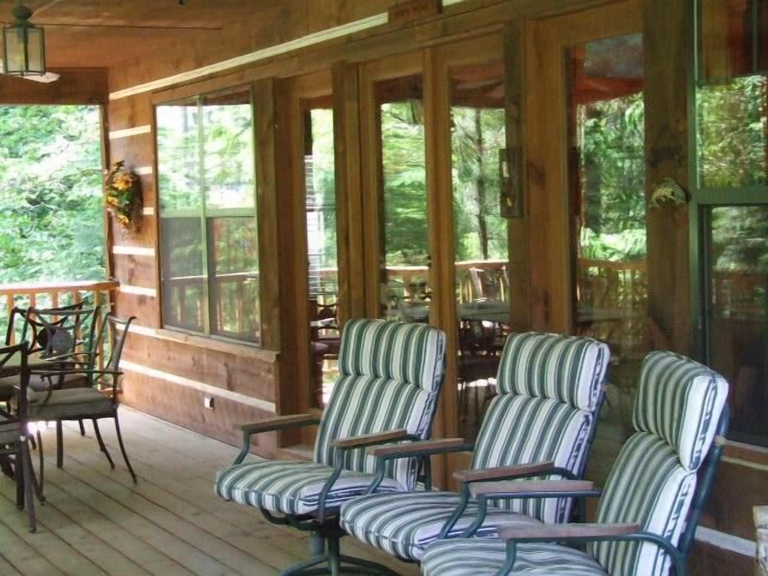 A wooden porch with striped armchairs and a table, large windows, and a view of greenery.