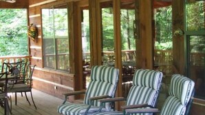 A wooden porch with striped armchairs and a table, large windows, and a view of greenery. - Pick's Riverside Retreat Nevaeh Cabin Rentals (Blue Ridge)