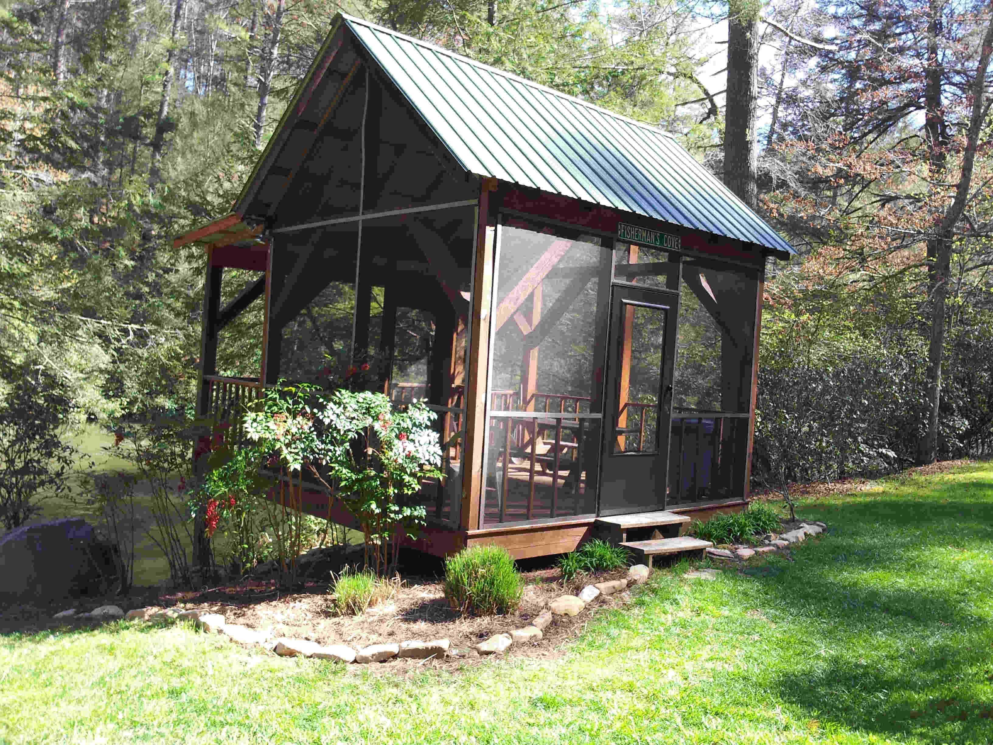 Una pequeña cabaña de madera con techo de metal, rodeada de un jardín y un estanque.