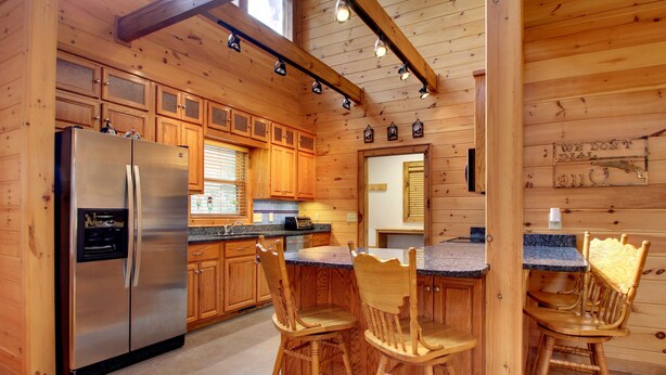 A wooden cabin-style kitchen with a stainless steel refrigerator, wooden cabinets, a granite countertop, and a dining area with wooden chairs.