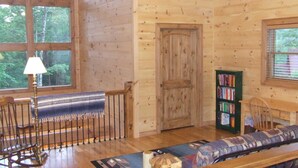 A cozy cabin interior with a wooden floor, a rocking chair, a sofa, a bookshelf, and a large window offering a view of the forest.