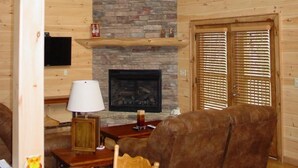A cozy cabin interior with a stone fireplace, wooden walls, a ceiling fan, and a large window.