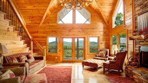 A log cabin interior with a high ceiling, a chandelier, multiple windows, and a fireplace.