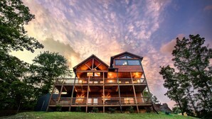 A two-story wooden cabin with a balcony, surrounded by trees and set against a cloudy sky.