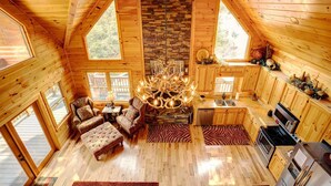 A log cabin interior with a central fireplace, wooden furniture, and large windows.