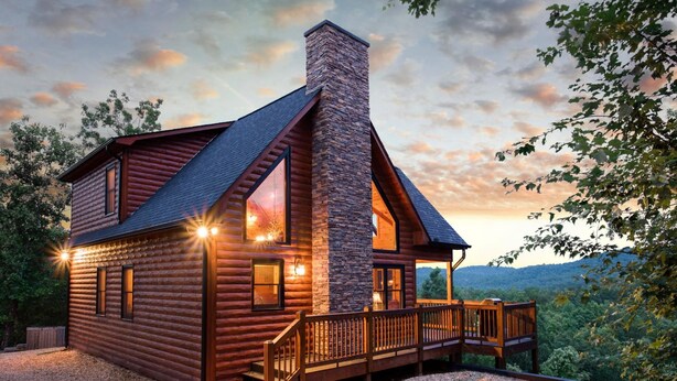 A log cabin with a stone chimney, large windows, and a wooden deck.