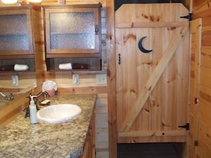 A wooden cabin bathroom with a granite countertop sink, a glass shower enclosure, and a wooden door with a crescent moon cutout.