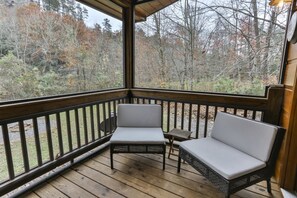 A covered porch with two wicker chairs and a small table, overlooking a forested area. - CAMP RISING FISH-NEVAEH CABIN RENTALS (Mineral Bluff)