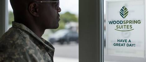 Una persona con uniforme militar de pie frente a una ventana con un cartel que dice "WOODSPRING SUITES HAVE A GREAT DAY!"