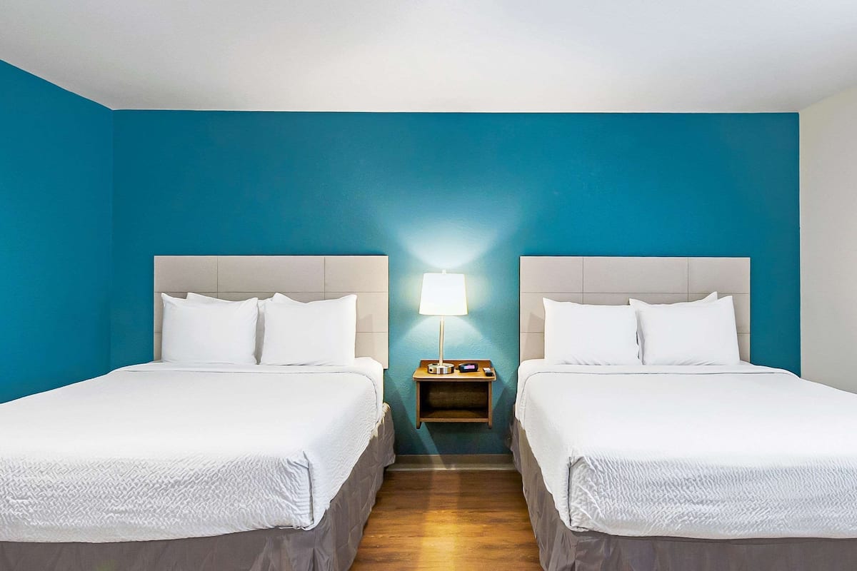 A hotel room with two beds, a nightstand with a lamp, and a blue accent wall.