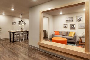 A modern living room with a grey sofa, orange ottoman, and a dining area with a wooden table and chairs. - WoodSpring Suites Washington DC Northeast Greenbelt (Greenbelt)