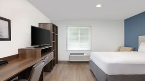 Una habitación de hotel con un escritorio de madera, televisor, cama con cubrecama gris y ventana con persianas.