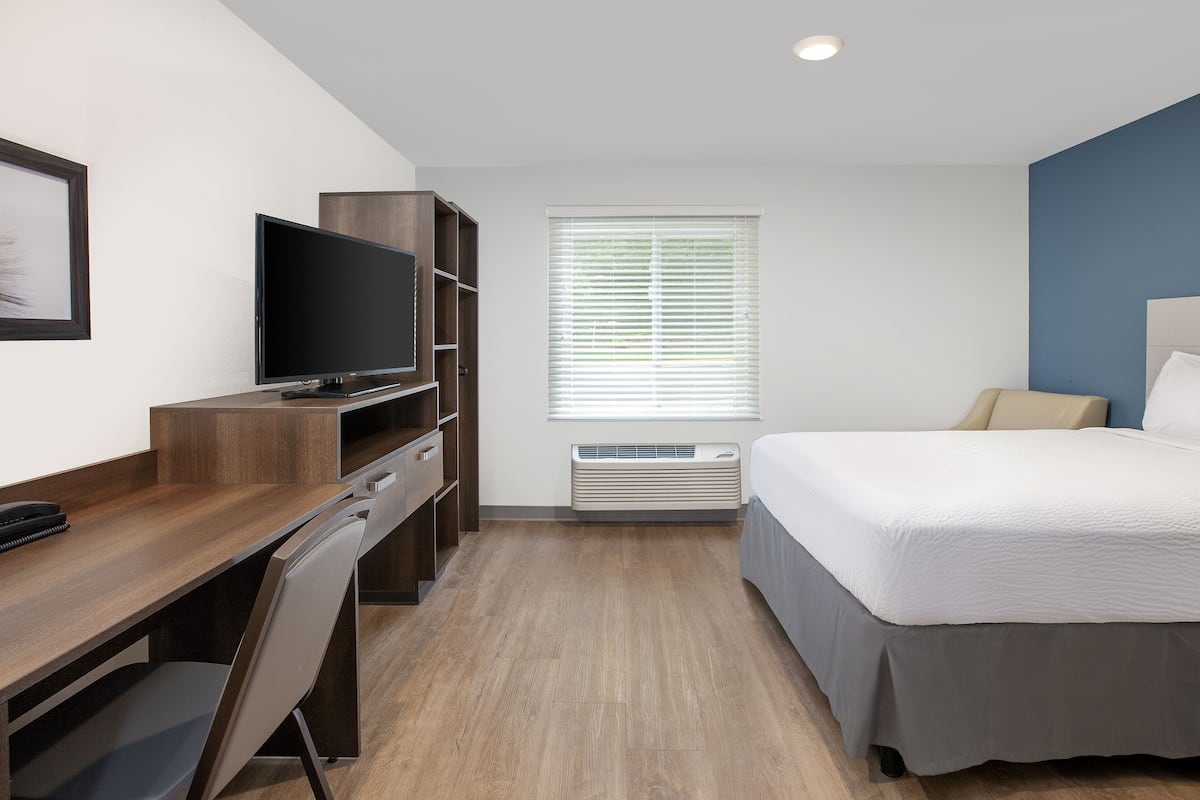 Een hotelkamer met een houten bureau, een televisie, een bed met een grijze plaid en een raam met jaloezieën.