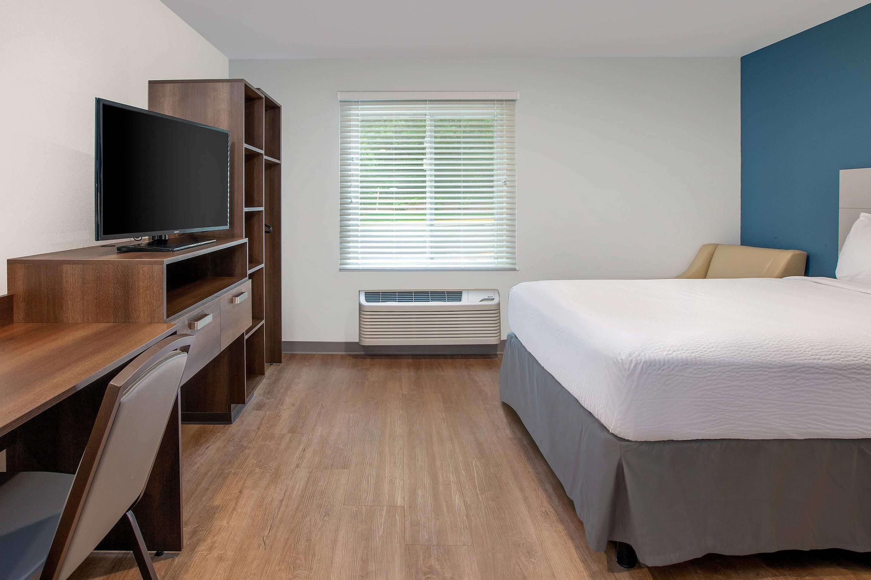 Habitación de hotel con escritorio de madera, televisor de pantalla plana, cama con colcha blanca, ventana con persianas y equipo de aire acondicionado.