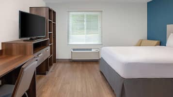 Quarto de hotel com escrivaninha de madeira, televisão de tela plana, cama com edredom branco, janela com persianas e aparelho de ar condicionado.