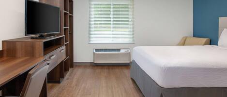 Habitación de hotel con escritorio de madera, televisor de pantalla plana, cama con colcha blanca, ventana con persianas y equipo de aire acondicionado.