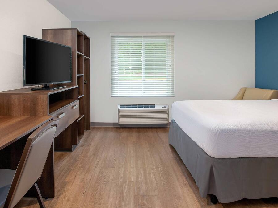 Quarto de hotel com escrivaninha de madeira, televisão de tela plana, cama com edredom branco, janela com persianas e aparelho de ar condicionado.