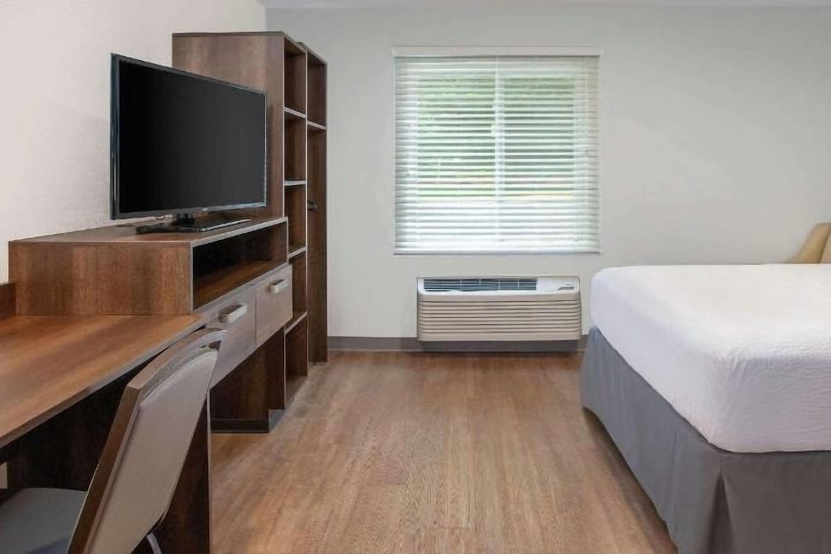 Een hotelkamer met een bed, een bureau met een televisie, een stoel, een raam met zonwering en een airconditioning.
