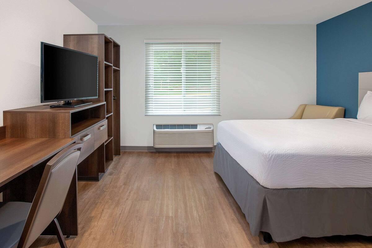 Hotelkamer met een houten bureau, een flatscreen televisie, een bed met grijze spreideken en een raam met zonwering.