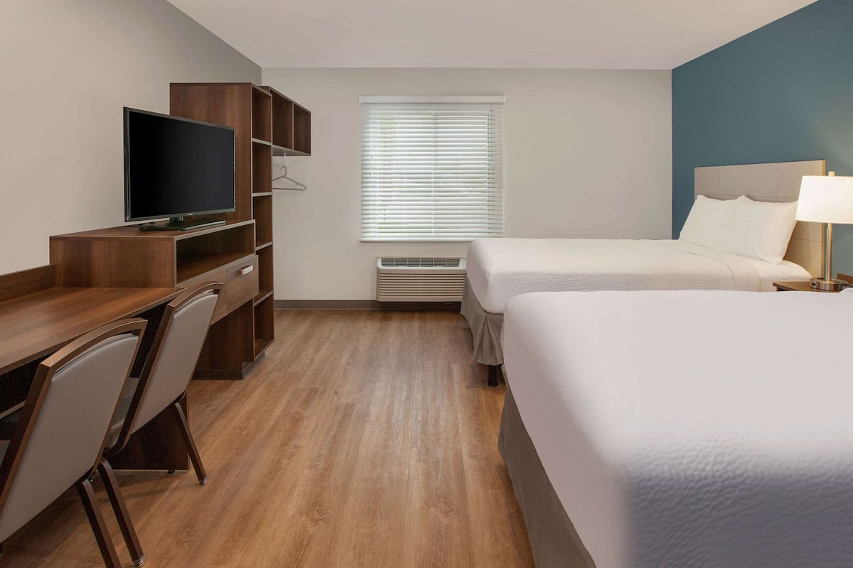 Een hotelkamer met een houten bureau, een televisie, een bed, een nachtkastje en een raam met jaloezieën.