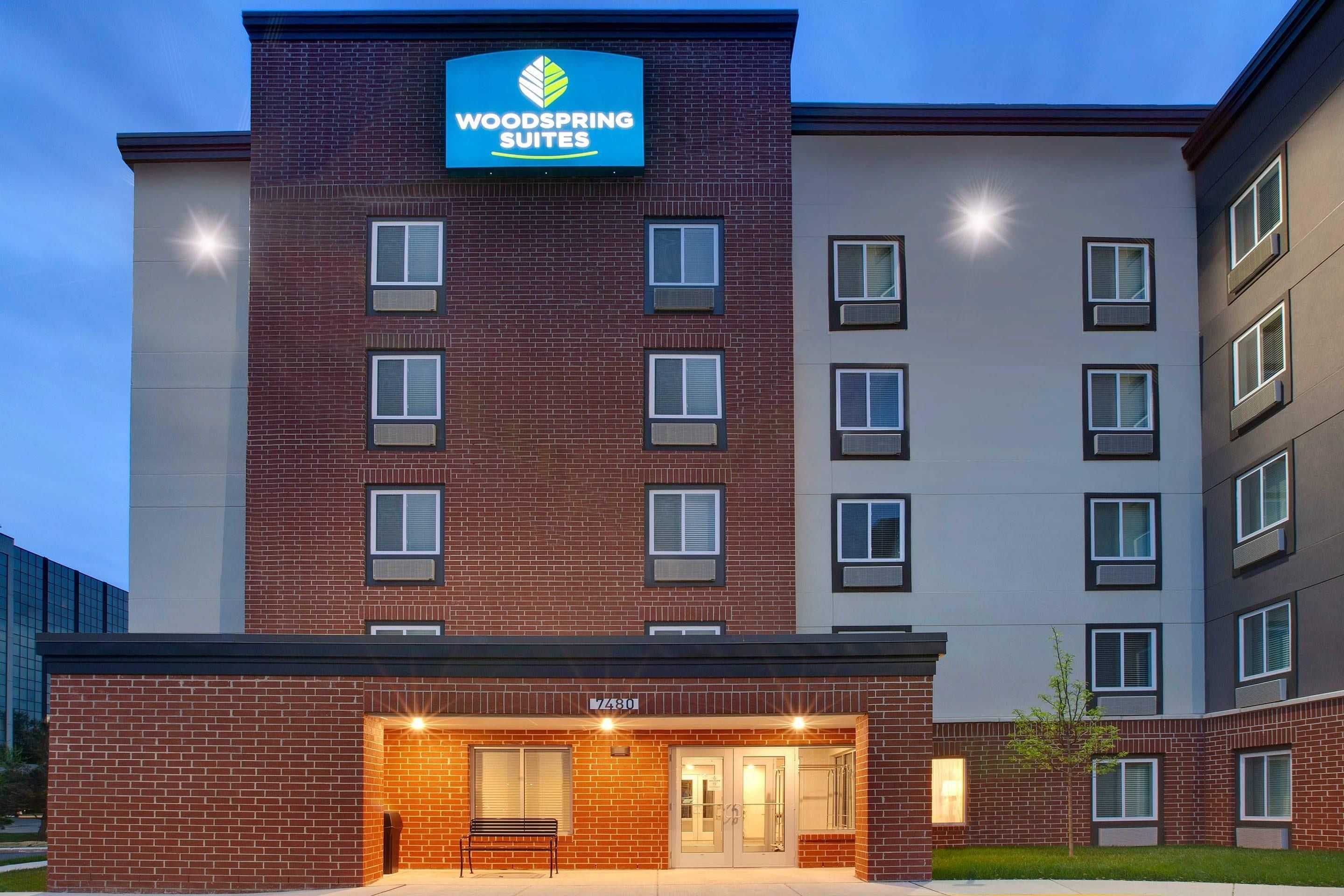 A multi-story hotel with a prominent sign for 'Woodspring Suites' and a ground-level entrance with a bench.