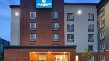 A multi-story hotel with a prominent sign for 'Woodspring Suites' and a ground-level entrance with a bench.