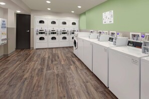 Une laverie équipée de plusieurs machines à laver et de plusieurs sèche-linge, d’un terminal de paiement et d’un panneau d’information fixé au mur.