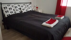 Een net opgemaakt bed met een zwart-wit gestreept kussen, een rood-wit dekbed en een klein nachtkastje met een rode vaas.