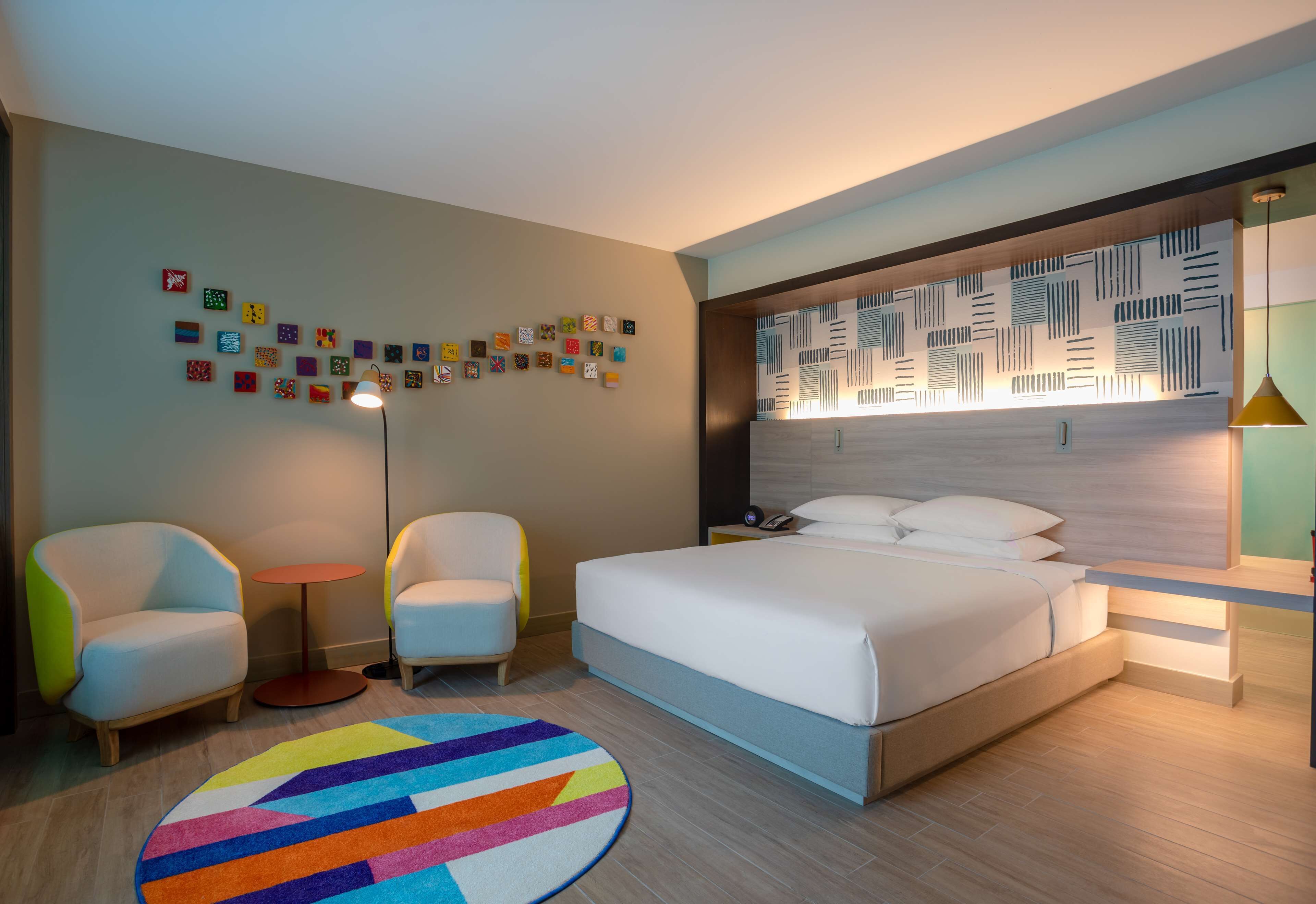A modern hotel room with a large bed, two armchairs, a small round table, a colorful rug, and a wall decorated with small art pieces.