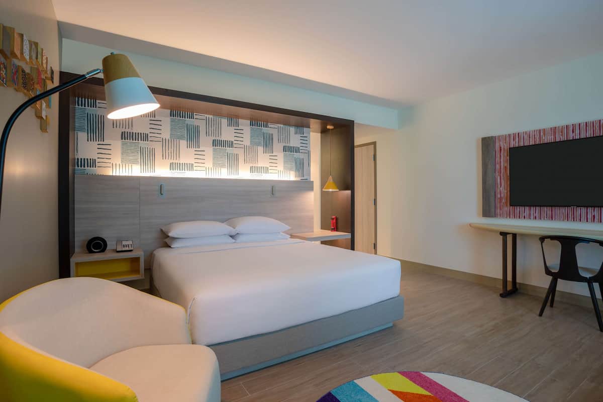 Une chambre d’hôtel moderne avec un grand lit, des tables de chevet, un bureau avec une chaise et un tapis coloré.