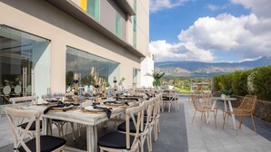 An outdoor dining area with multiple tables and chairs, a modern building with large windows, and a mountainous landscape in the background. - Hyatt Centric San Salvador (Antiguo Cuscatlán)