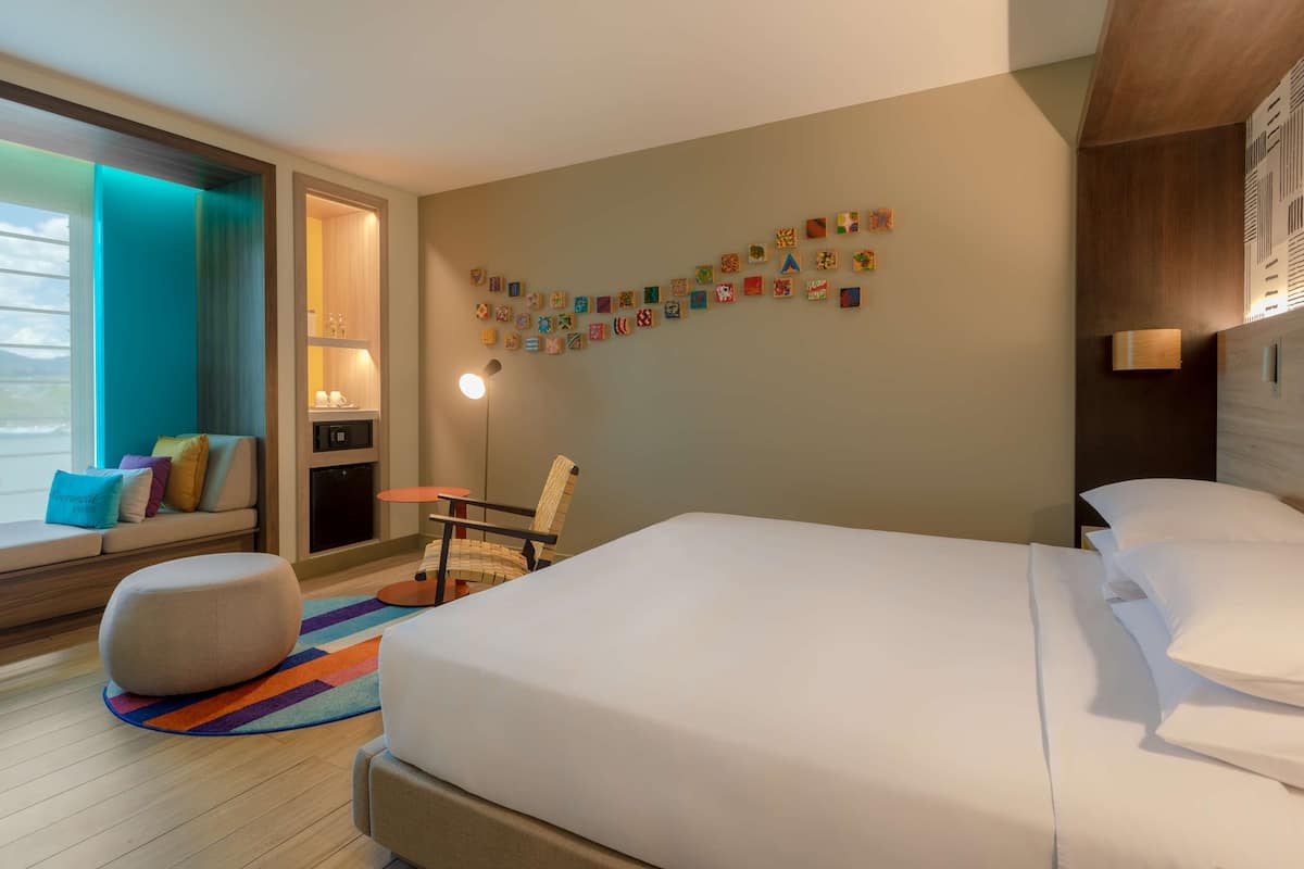 Une chambre d’hôtel moderne avec un grand lit, un coin salon comprenant un canapé et un pouf, une petite table, une chaise et un mur décoré de cartes postales colorées.