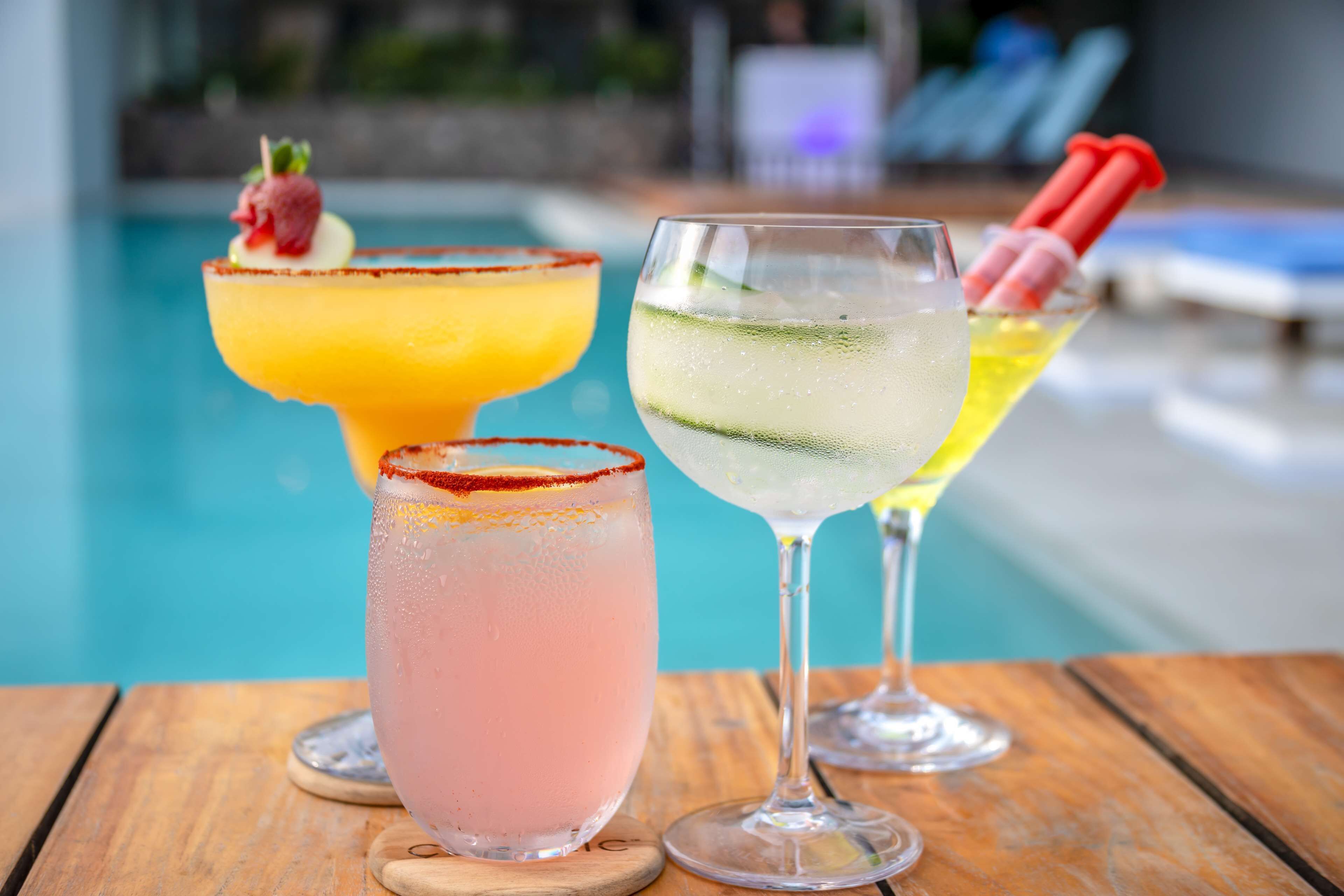Three cocktails on a wooden table by a poolside.