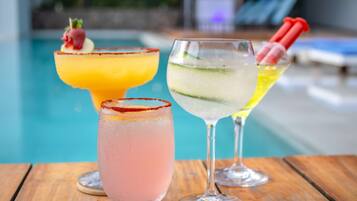 Three cocktails on a wooden table by a poolside.
