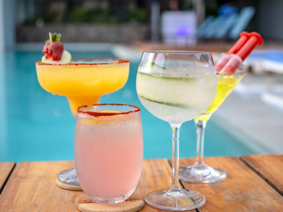 Three cocktails on a wooden table by a poolside.