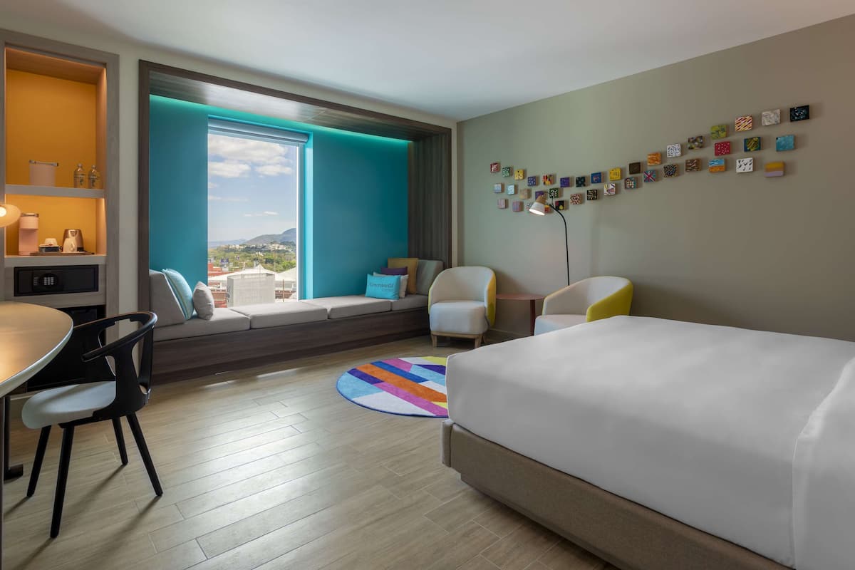 Une chambre d’hôtel avec un lit, un bureau, une chaise, un canapé, un tapis coloré et une vue sur la ville.