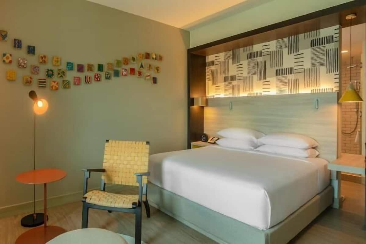 Une chambre d’hôtel comprenant un lit, une chaise, une petite table, une lampe et un mur orné de divers objets décoratifs.