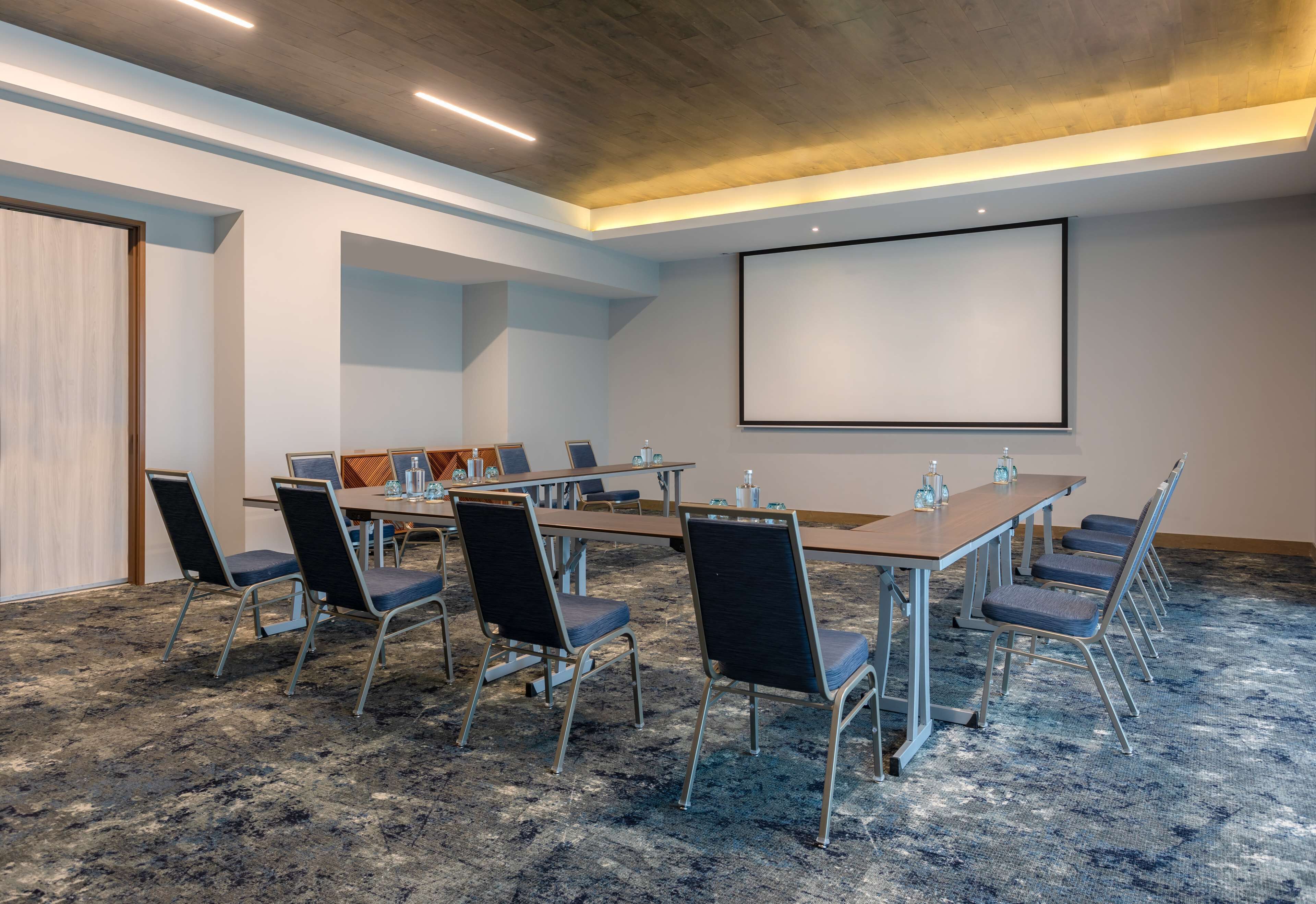 A conference room with a long table, chairs, a projector screen, and a carpeted floor.