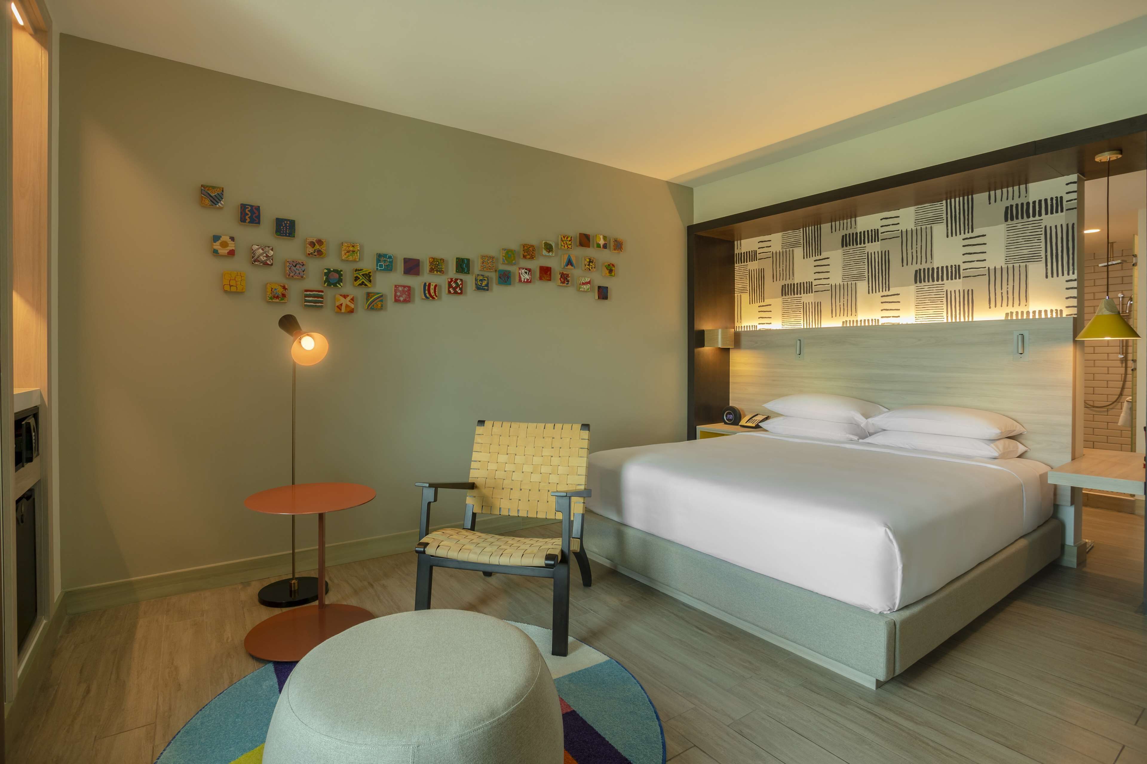 Quarto de hotel com cama, uma cadeira, uma mesinha e um detalhe decorativo na parede.