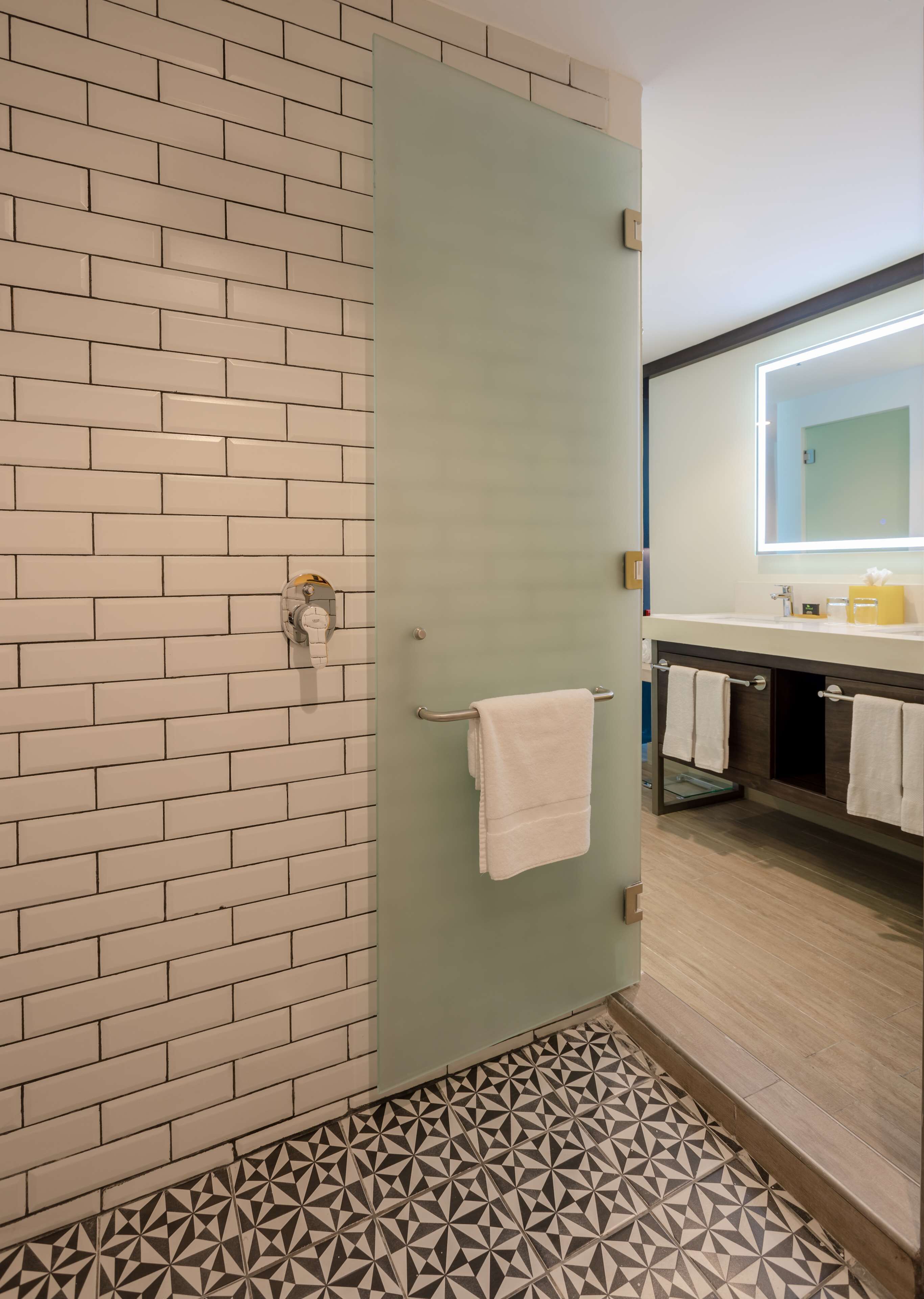 A bathroom with a tiled wall, a glass door, a patterned floor, and a mirror with a light.