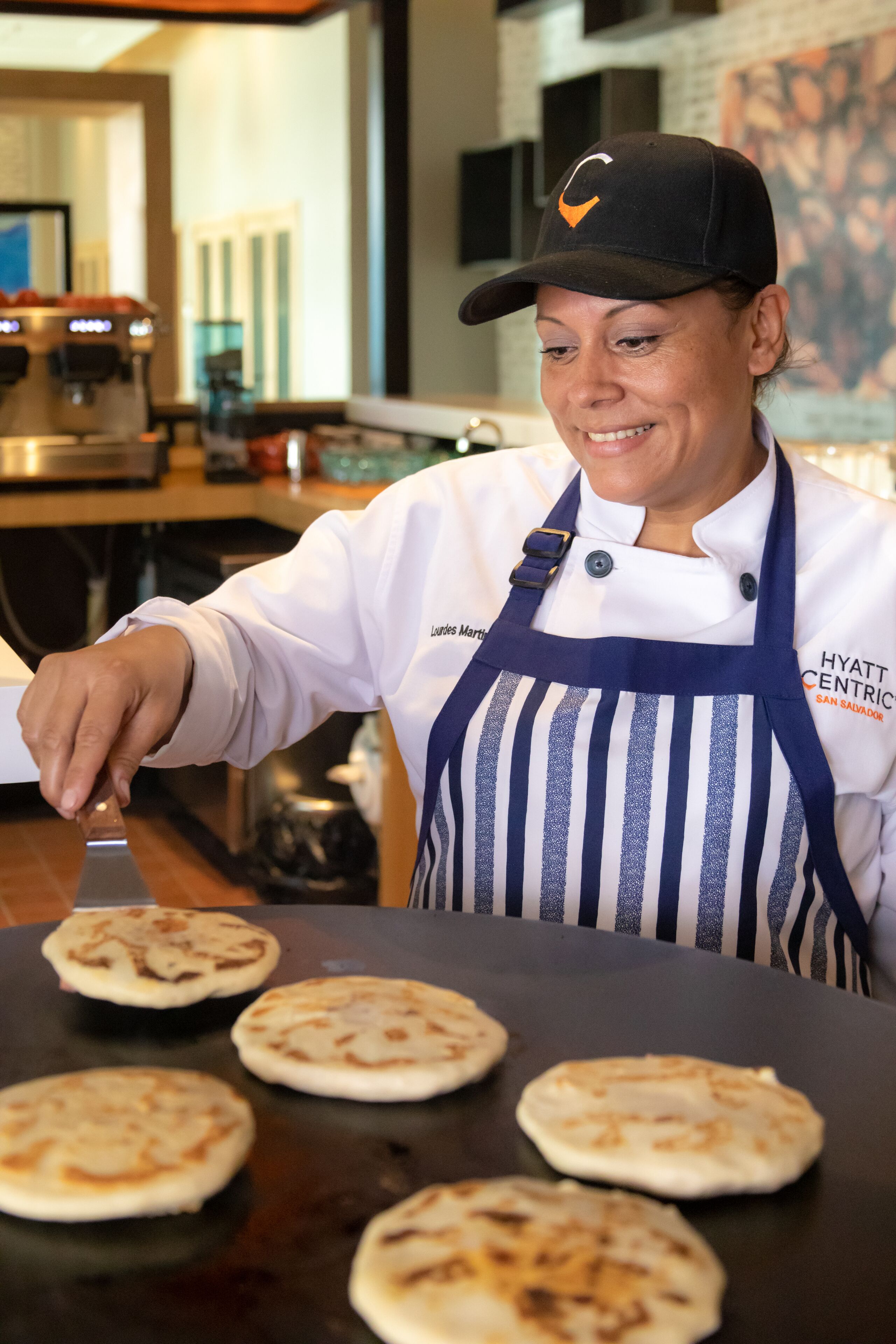 A person in a white chef's coat and striped apron cooking flatbreads on a griddle.