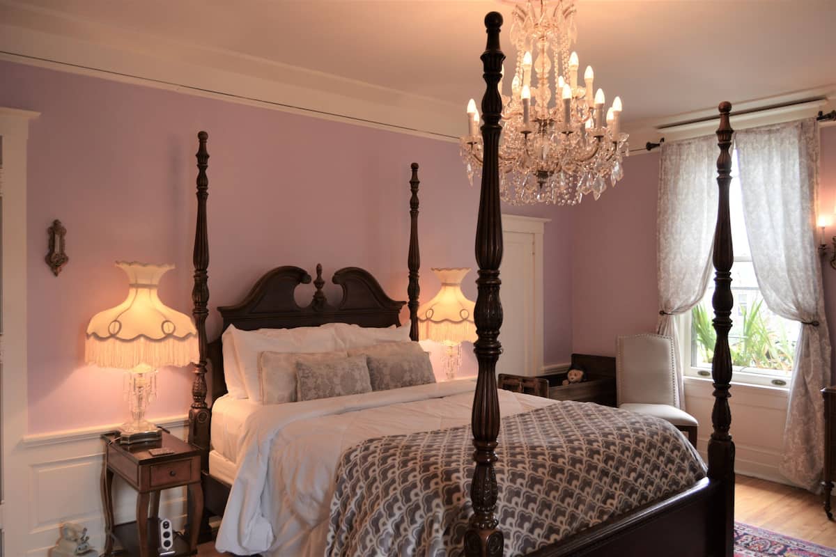 A four-poster bed with a chandelier, a bedside table with a lamp, and a window with curtains.