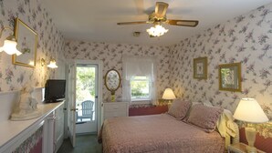 A bedroom with floral wallpaper, a bed with pink bedding, a ceiling fan, and a desk with a television.