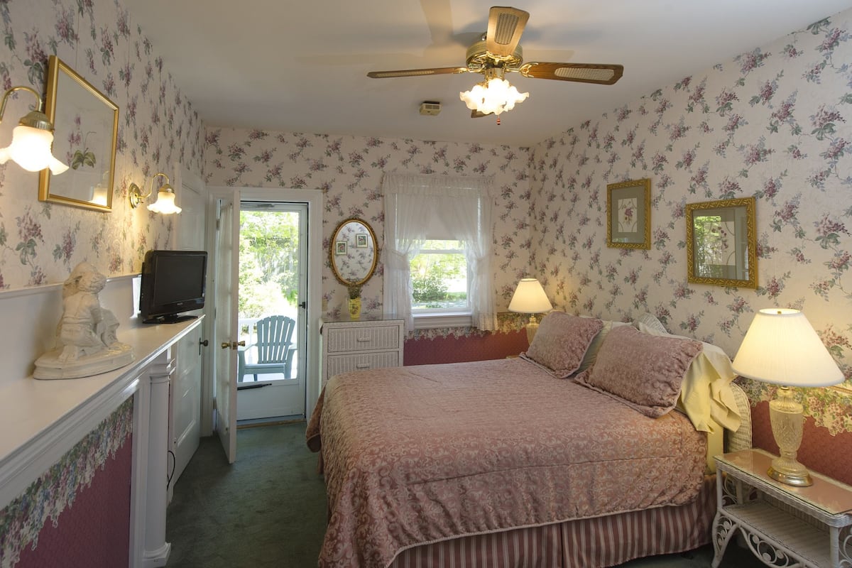 A bedroom with floral wallpaper, a bed with pink bedding, a ceiling fan, and a desk with a television.