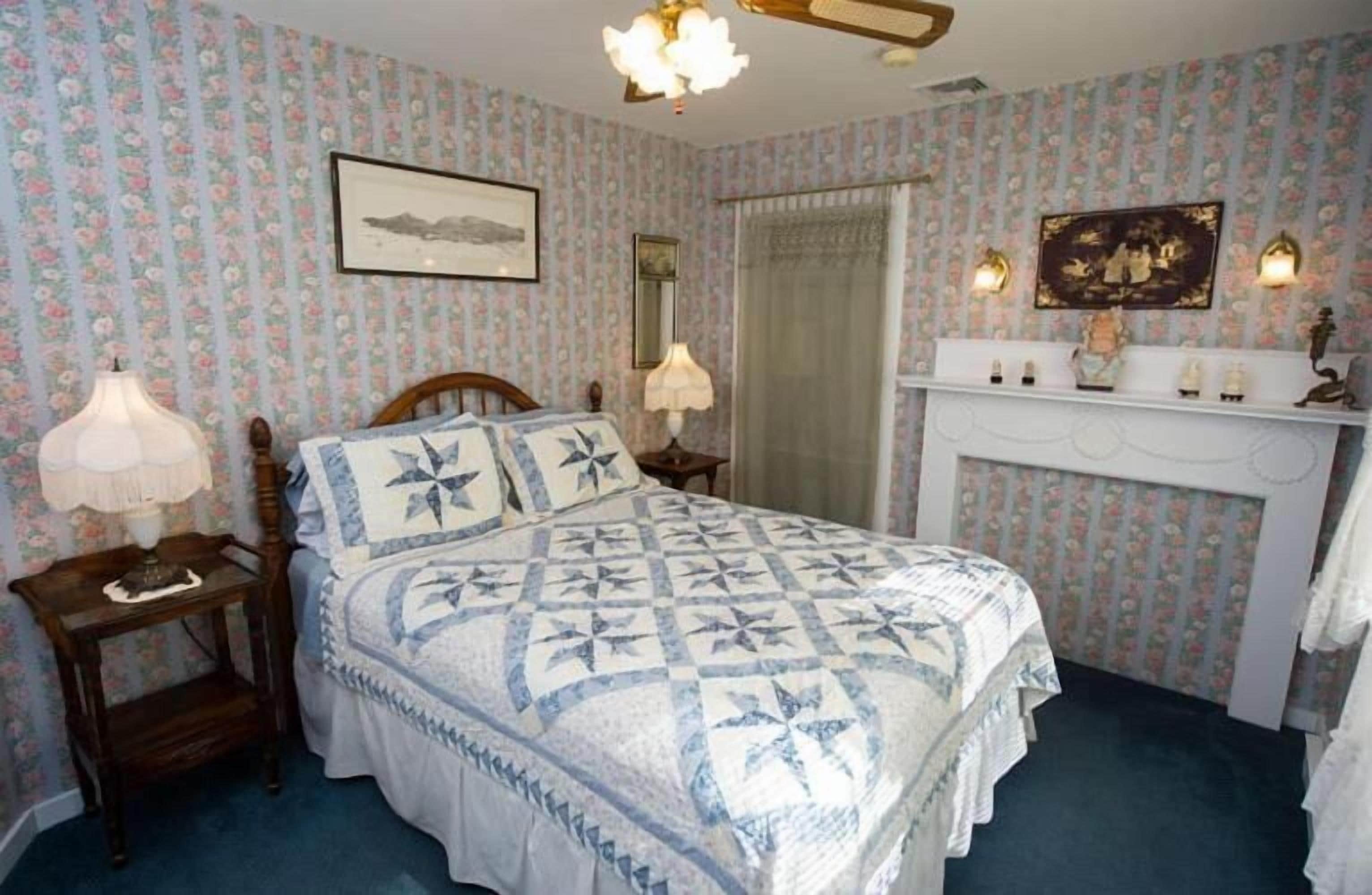 A bedroom with floral wallpaper, a bed with a quilt, a dresser with a mirror, and wall sconces.