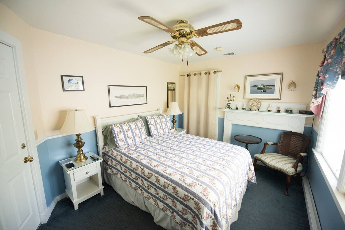 A bedroom with a bed, nightstand, chair, ceiling fan, and wall decorations.