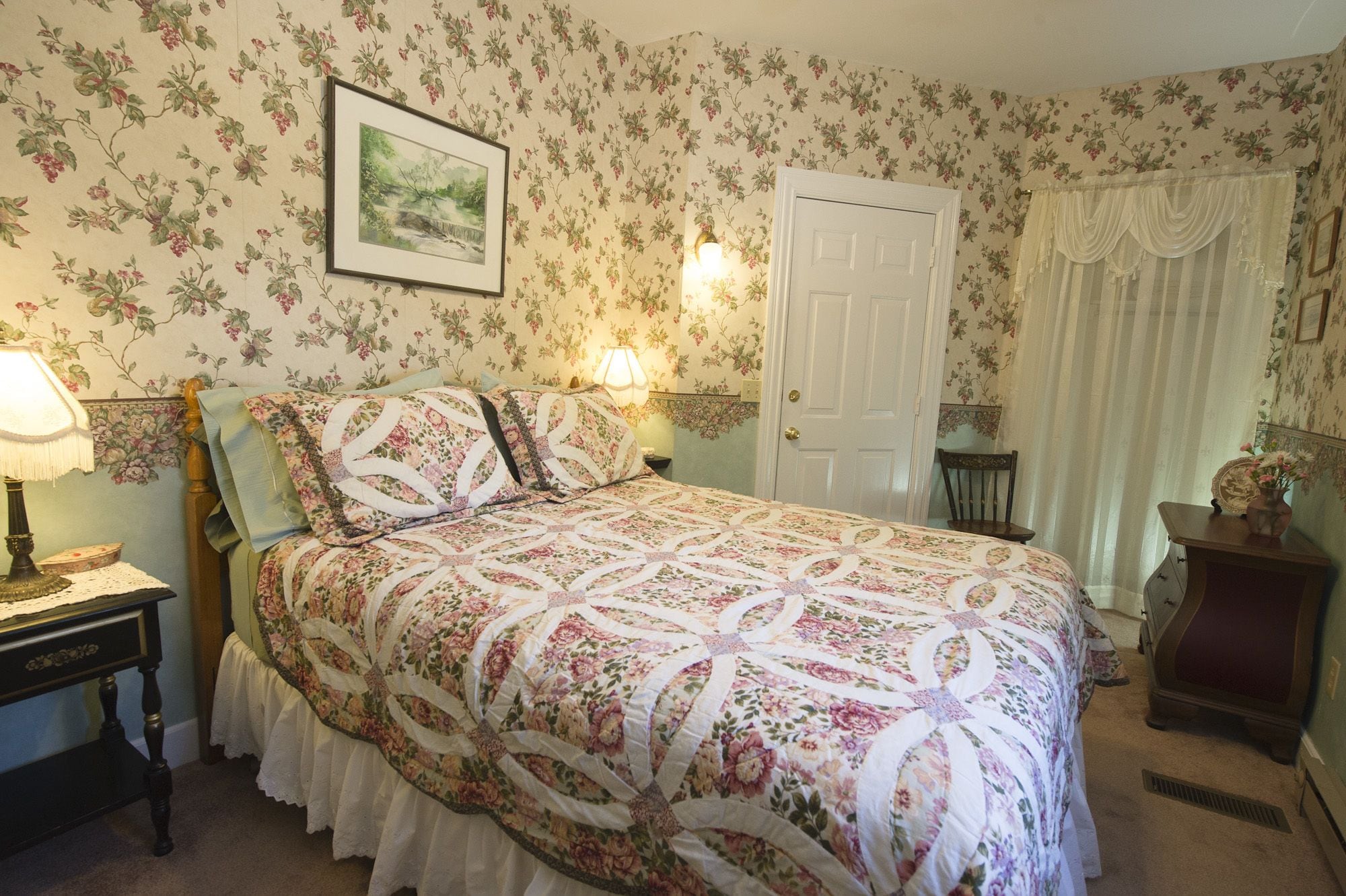 A bedroom with floral wallpaper, a bed with a floral quilt, a nightstand with a lamp, a door, and a window with curtains.